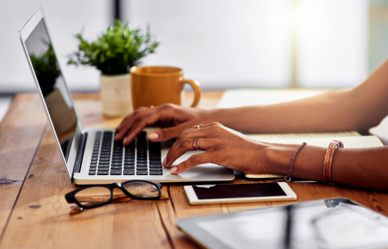 Online-Bewerbung leicht gemacht Frauenhände tippen auf Laptop, daneben auf dem Tisch ein Tablet, Brille, Handy, Kaffeetasse und Notizbuch
