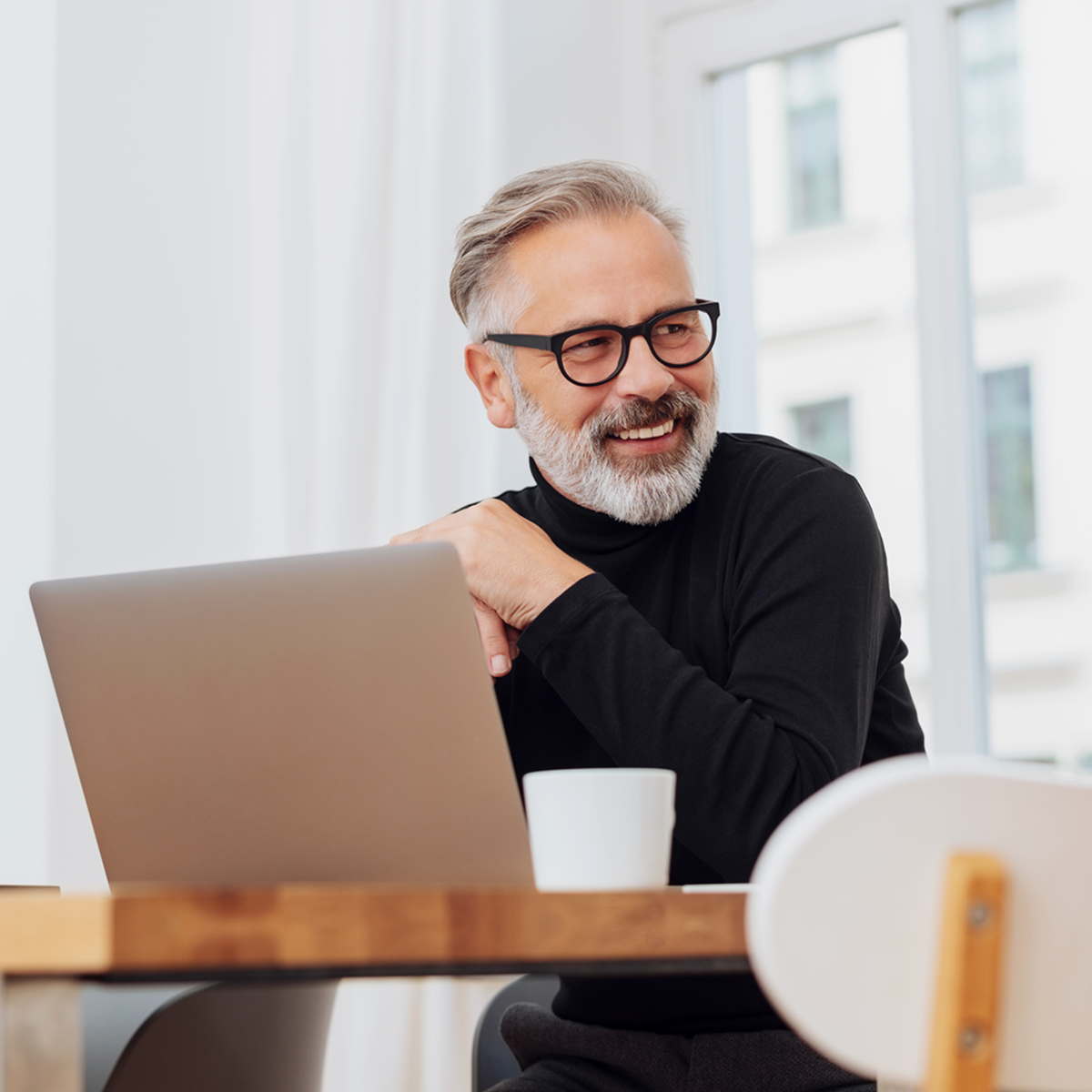 Grauhaariger Mann mit Vollbart und Brille sitzt vor Laptop und lächelt
