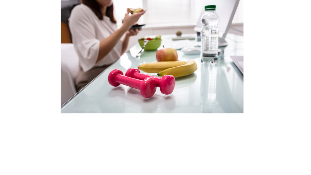 Zwei Kurzhanteln, zwei Bananen und ein Apfel liegen auf Schreibtisch, im Hintergrund frühstückt eine Frau vorm Computer