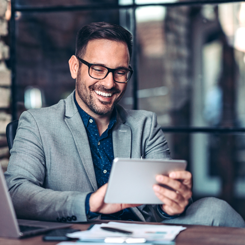 Lachender Mann mit Brille sitzt mit Touchpad am Schreibtisch