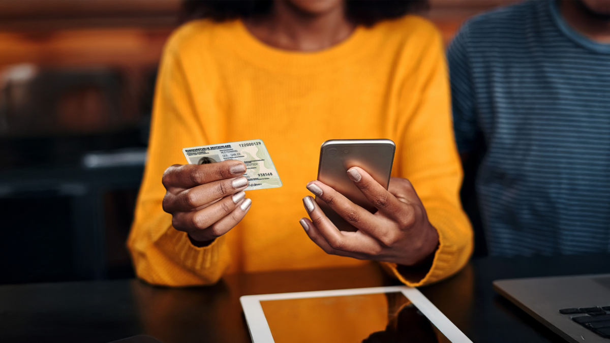 Frau in orangenem Pullover haelt Ausweis und Smartphone in der Hand