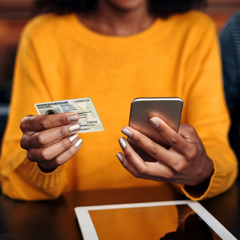 Frau in orangenem Pullover haelt Ausweis und Smartphone in der Hand